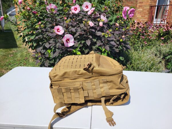 Side of a tan backpack on a white table with flower bush in the background