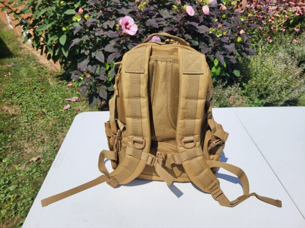 Back of a tan backpack on a white table with flower bush in the background