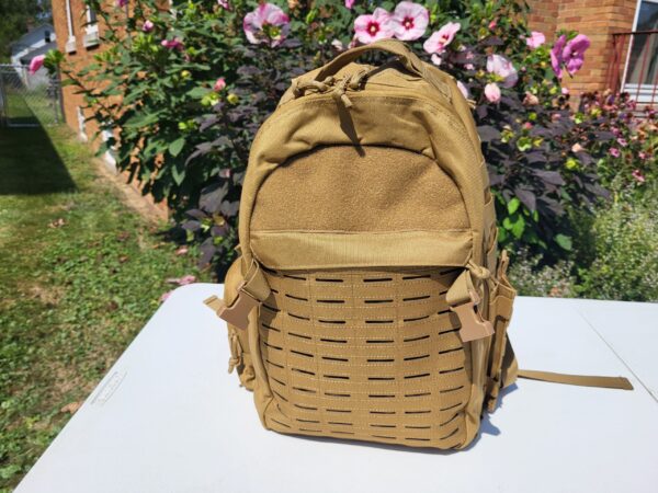 Front of a tan backpack on a white table with flower bush in the background