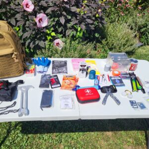 Tan backpack with multi tool, hammer, rope, glow sticks, tooth paste, tooth brush and related items on a table.