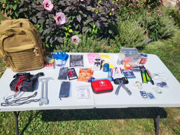 Tan backpack with multi tool, hammer, rope, glow sticks, tooth paste, tooth brush and related items on a table.